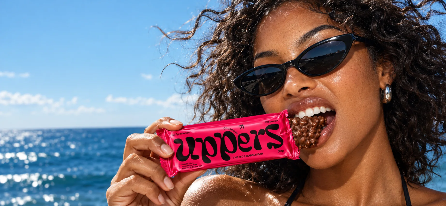A woman in cat-eye sunglasses bites into an opened Uppers bar on a sunny beach, ocean behind her