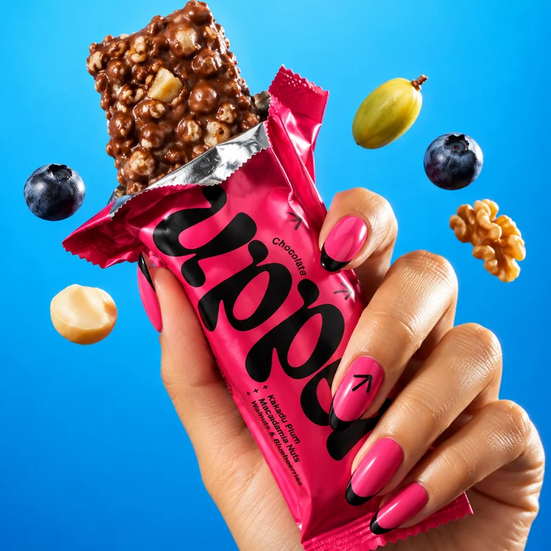 A hand with pink and black nails holding an opened Uppers bar, with a blueberry, Kakadu plum, walnut and macadamia floating around it on a blue backdrop